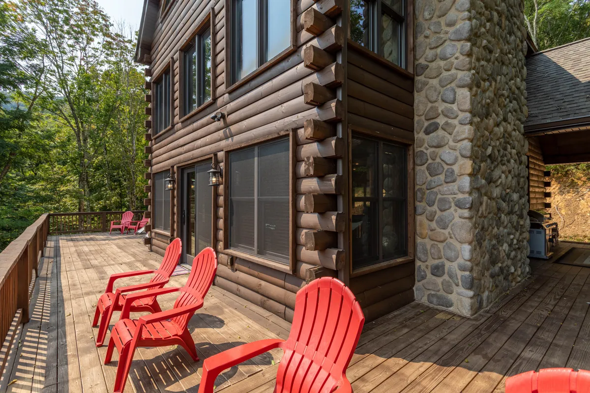 A welcoming wooden deck with red Adirondack chairs set beside a stone wall and large windows. The tranquil outdoor setting is ideal for morning coffee, stargazing, or simply relaxing while surrounded by nature's serenity.