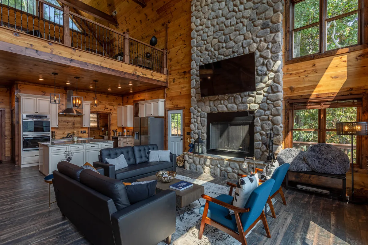 An open-concept living area with a grand stone fireplace, exposed wooden beams, comfortable seating, and a clear view into the kitchen. The large windows bathe the space in natural light, highlighting both the cozy living room and the functional, chef-friendly kitchen setup.
