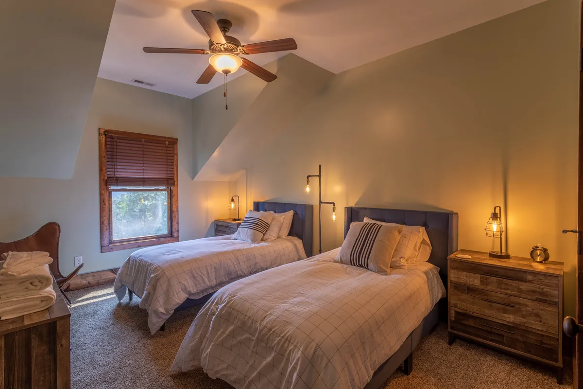A cozy bedroom with twin beds placed side by side, each with its own nightstand and a window letting in soft natural light. The simple, soothing decor suggests a comfortable sleep environment for siblings or friends.