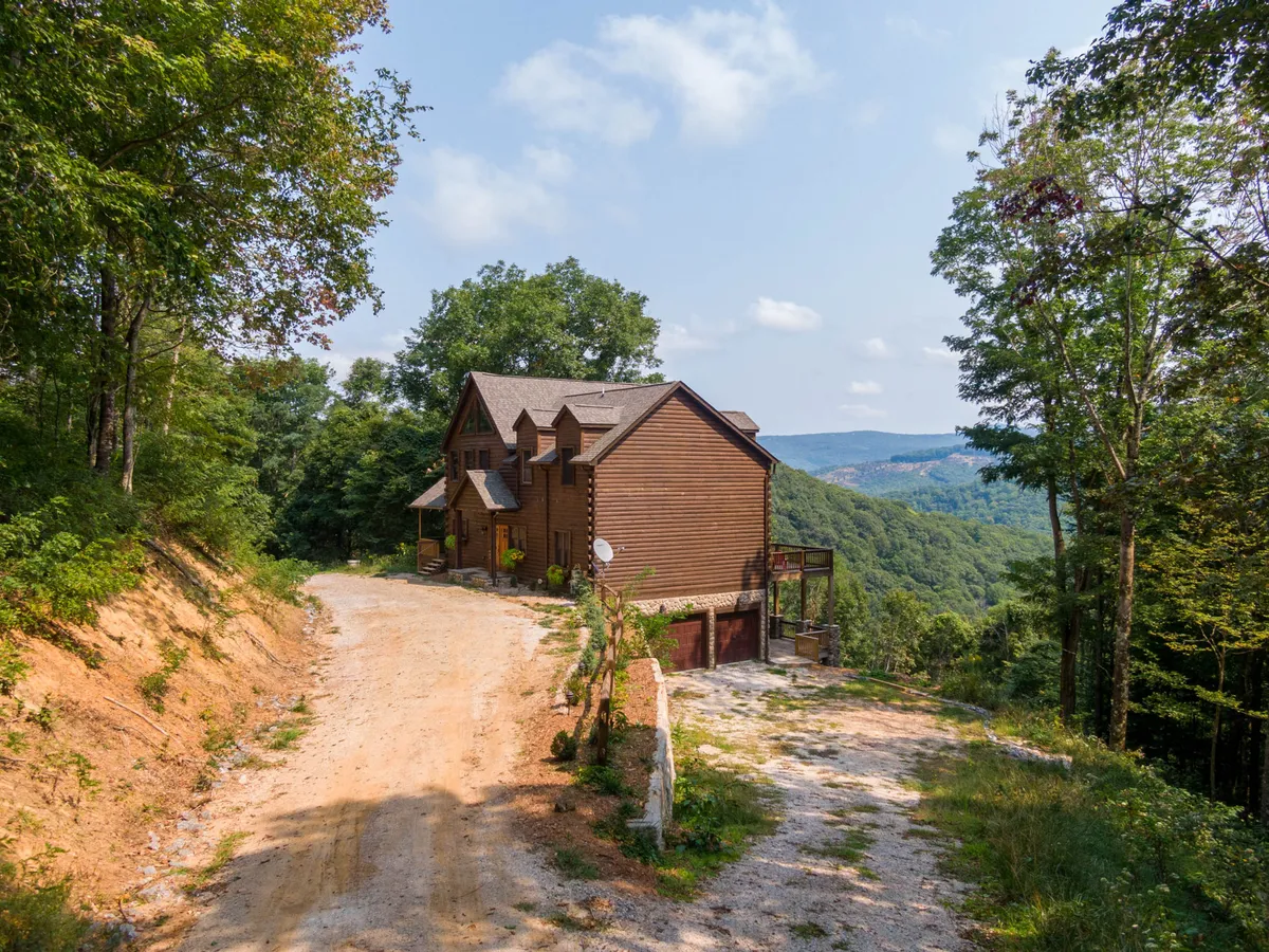 A peaceful exterior shot of the lodge featuring a wooden house, a gravel driveway, and a backdrop of mountain views and surrounding trees. The home sits nestled in nature, suggesting immediate access to the outdoors.