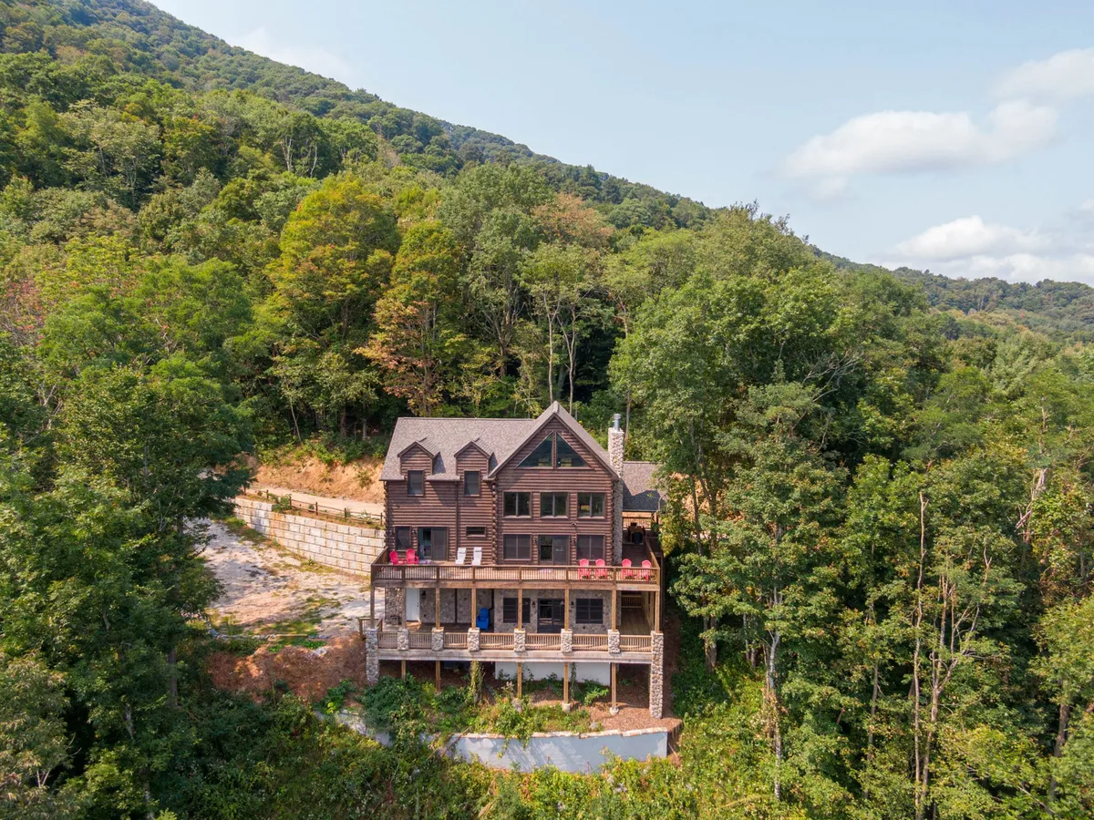 A wide exterior view of the wooden lodge nestled among mature trees, with a spacious porch featuring seating areas and a stunning mountain backdrop. The tranquil setting emphasizes the lodge's seclusion, privacy, and natural surroundings.