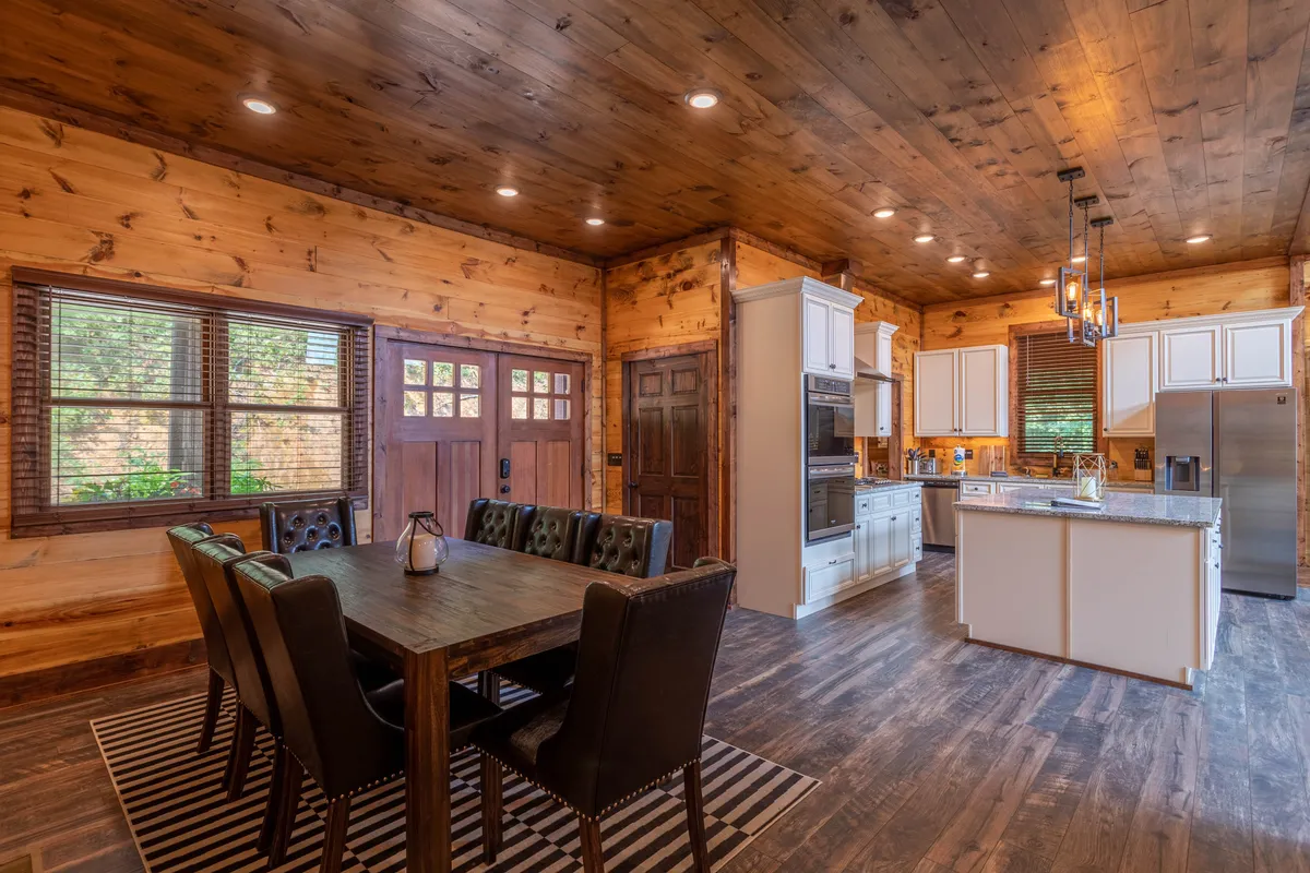 This image centers on the communal dining table, kitchen island, and wooden cabinets, all illuminated by natural light streaming through expansive windows. The space exudes warmth and sociability, with the kitchen and dining areas seamlessly flowing together—perfect for family meals, entertaining, and connecting over food.