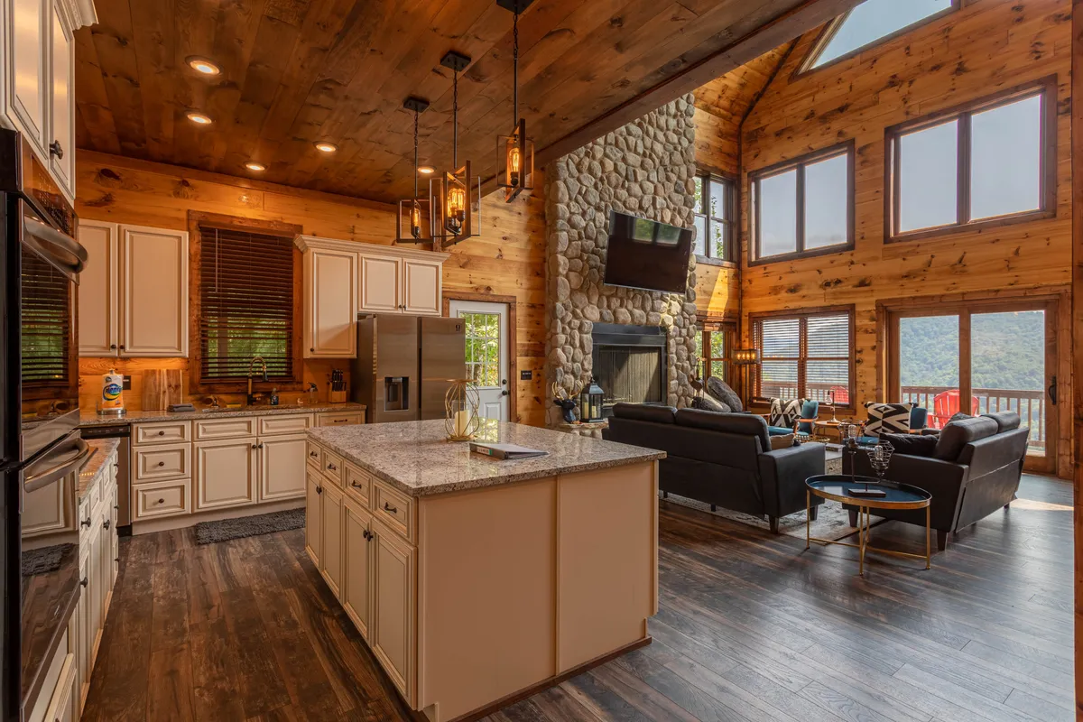 A broad view of the great room showing the large kitchen island, stone fireplace, generous windows framing mountain vistas, and a plush seating area, all arranged for easy conversation and relaxation.
