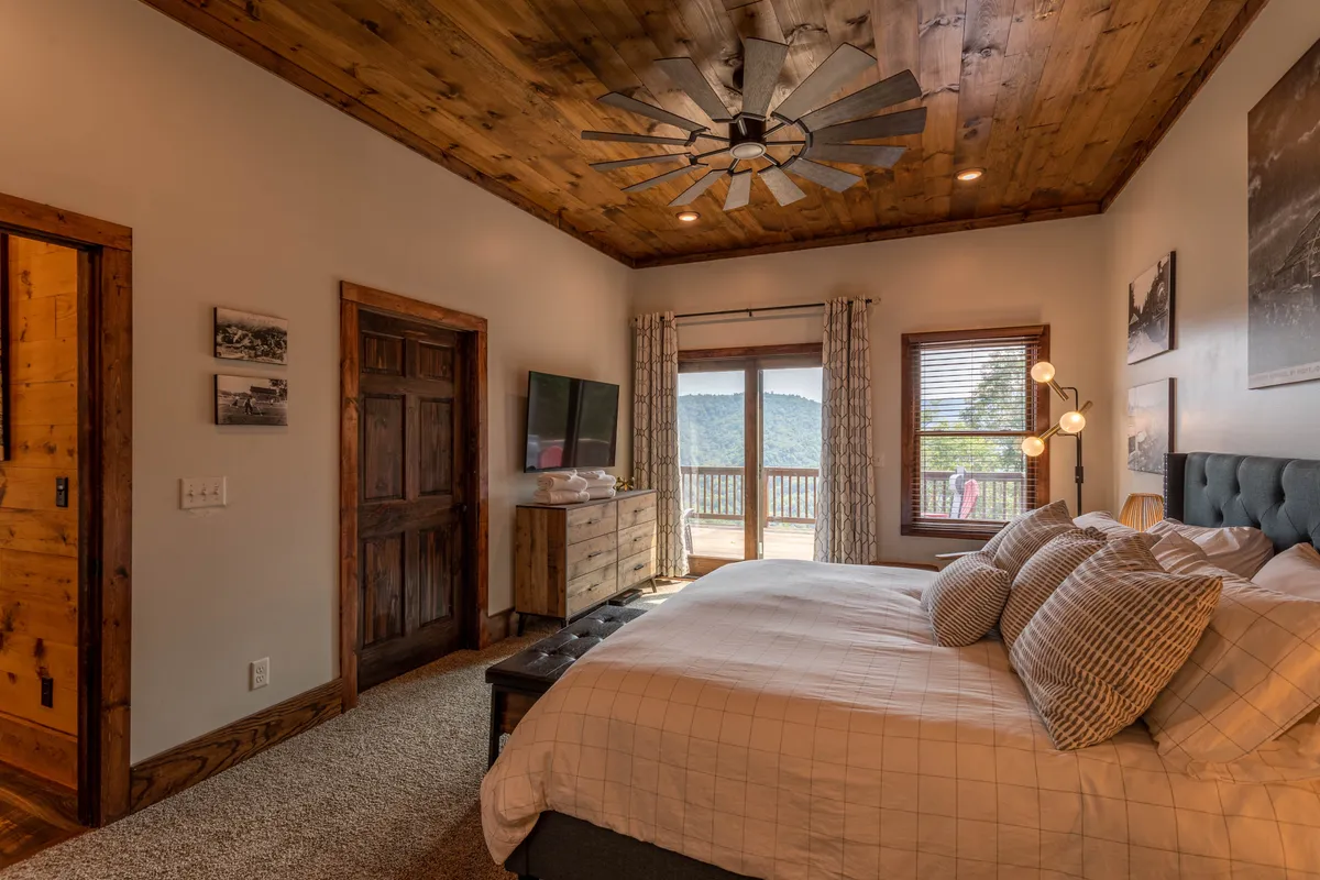 A king-sized bed with decorative pillows is set against wooden elements, complemented by a large window offering scenic views and a ceiling fan above. A nightstand with a lamp sits beside the bed, and natural light fills the room, creating a peaceful and inviting master suite atmosphere.