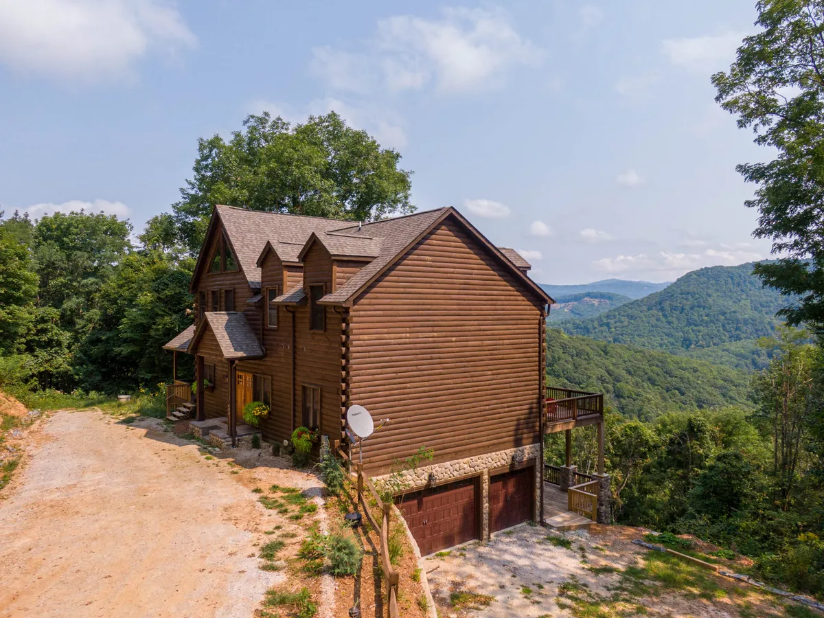The image shows a spacious wooden cabin set against a stunning backdrop of rolling North Carolina mountains. The cabin sits privately with no neighboring homes in sight, and features a broad deck perfect for relaxing or taking in the sweeping views. The driveway offers easy access, while the open yard invites guests to gather around for evening bonfires or enjoy tranquil mornings outdoors.
